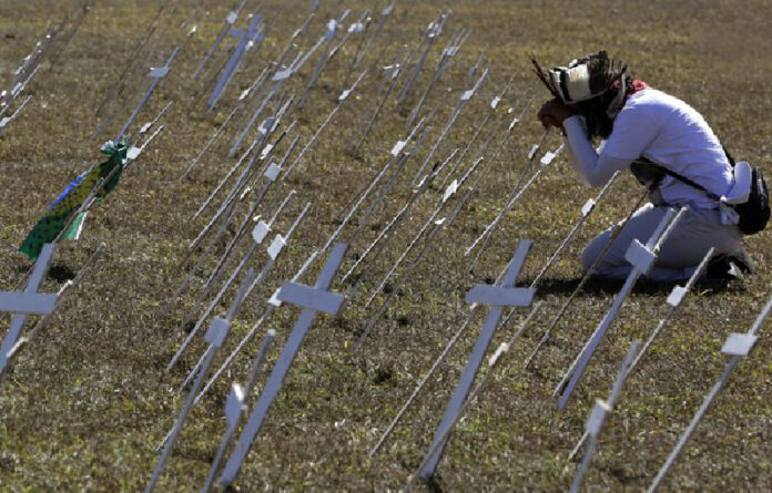 Un manifestante se arrodilla ante cruces que representan los miles de muertos por coronavirus, frente al Congreso de Brasil, en Brasilia. Foto Ap.